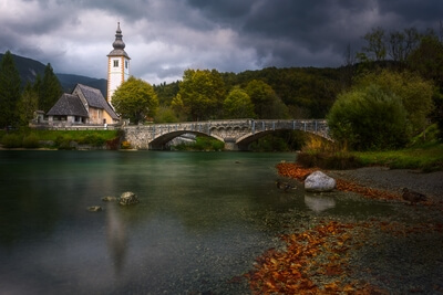 Even with the ominous clouds there was something idyllic about Lake Bohinj that day. Out of the 3 compositions I shot that day this was my favourite and thats why I took my time to post process this image. Two exposures blended - a longer exposure to smooth out the water and more details in the shadows and the normal exposure which in this case was the darker of the two exposures for the sky.

Loved the autumn leaves lining up the shore leading the eye to the quaint old bridge and to the chapel.
