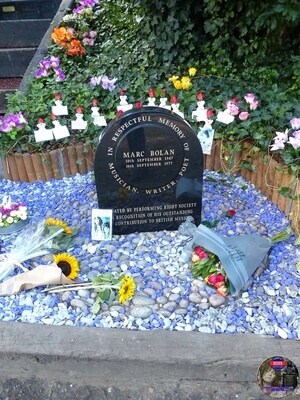 Flowers by the PRS Memorial at Marc Bolan's Rock Shrine, Gipsy Lane, Barnes, SW15 5RG London. Photograph taken on the 16th September 2021 - The 44th Anniversary of his passing at this site. Marc's Shrine has been cared for since 1999 by TAG (T.Rex Action Group) via an in-perpetuity lease on the site. The PRS Memorial was installed in September 1997 for the 20th Anniversary. Five years later the bronze bust of Marc Bolan was installed for the 25th Anniversary. Both were unveiled by Marc Bolan's only heir Rolan Bolan who was just 10 days short of his second birthday when his dad died. That fateful night Rolan was asleep at his paternal grandparents home in Putney. Marc and his partner, Rolan's mother were on their way home at 142 Upper Richmond Road West, Sheen.  