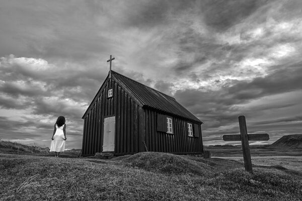 Small black isolated church with two marked graves out front, situated overlooking open grasses across from Elderberg mountain in Iceland. 