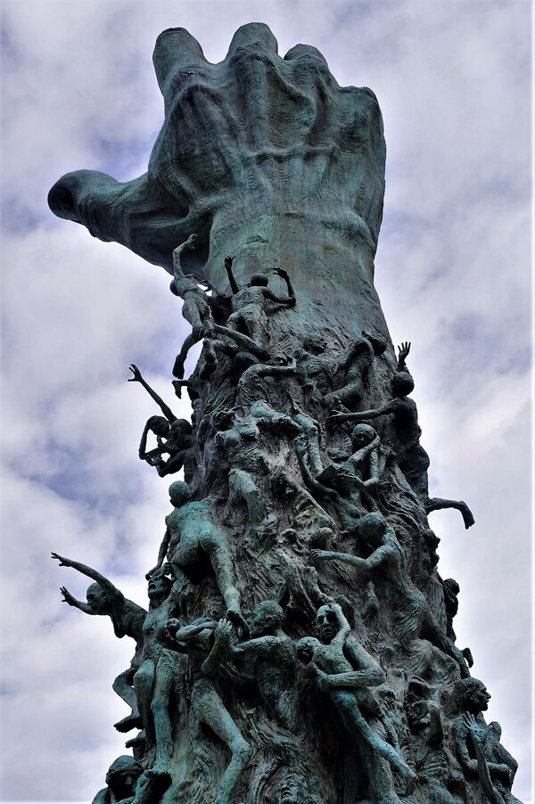 Portrait image, in which an arm is raised, fist extended to the sky.The arm is wrapped with figures of people ascending or falling.The 'dark' shade of the sculpture contrast with the 'light' of the sky. 